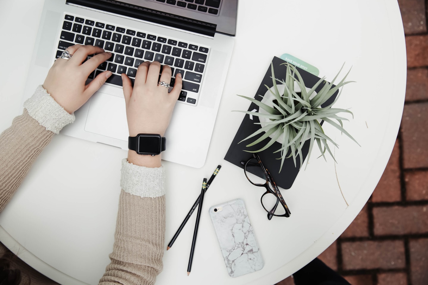 Top-down view of a person typing on a laptop at a white round table, with a smartwatch, pencils, glasses, and a plant nearby on the desk.
